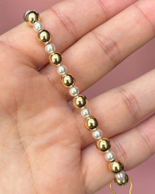 Gold and pearl beaded bracelet on a hand against a pink background