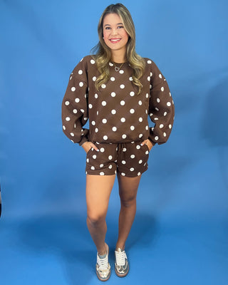 A woman modeling a brown and white polka dot sweatshirt and matching shorts set against a solid blue studio backdrop.