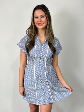 Smiling woman standing against a neutral background wearing a blue floral short-sleeve mini dress with button-front detail and contrast trim, styled with nude heeled sandals.