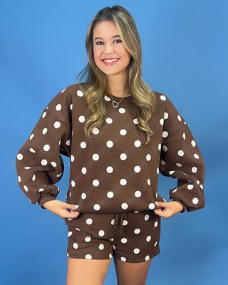 A woman modeling a brown two-piece lounge set with white polka dots, consisting of a long-sleeve sweatshirt and matching shorts, against a solid blue background.