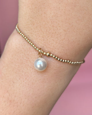 Gold bracelet with a pearl charm on a wrist against a pink background