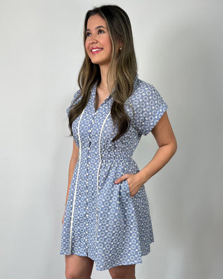 Smiling woman standing against a neutral background wearing a blue floral short-sleeve mini dress with button-front detail and contrast trim, styled with nude heeled sandals.