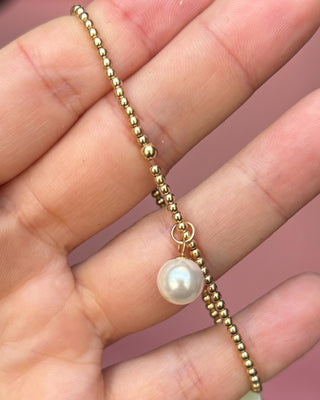Gold beaded chain with a pearl pendant held between fingers against a pink background