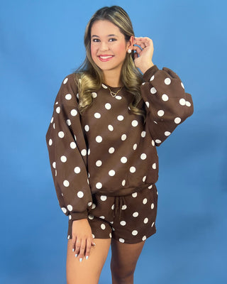 A woman modeling a brown and white polka dot lounge set consisting of a long-sleeve sweatshirt and matching shorts, posed against a solid blue studio background.