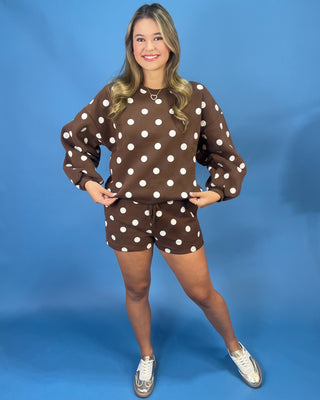 A woman modeling a brown and white polka dot sweatshirt and matching shorts set against a solid blue studio backdrop.