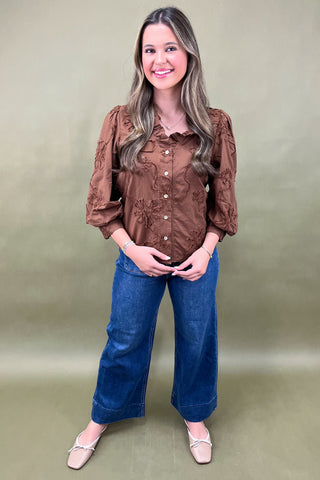 Woman wearing a brown blouse with floral patterns and blue jeans against a plain background