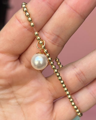 Gold beaded necklace with a pearl pendant held between fingers against a pink background