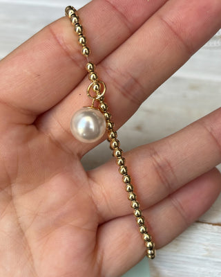 Gold beaded bracelet with a pearl held in a hand against a neutral background