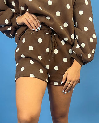 A person wearing a brown and white polka dot lounge set, featuring a long-sleeve top and matching shorts with a drawstring waist, posed against a solid blue background.