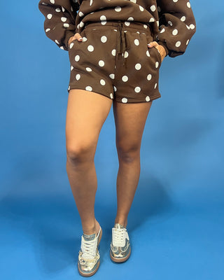 A person wearing brown and white polka dot sweatshirt and shorts set, paired with distressed white sneakers, standing against a solid blue studio background.