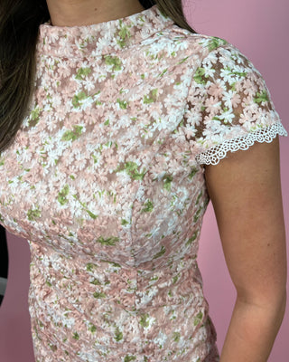 Blush pink floral midi dress with delicate lace trim, featuring a fitted bodice, short flutter sleeves, and a softly flared skirt; styled with gold strappy heels and minimal jewelry against a pink backdrop.