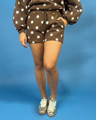 A person wearing a brown and white polka dot sweatshirt and matching shorts, paired with metallic silver sneakers, standing against a solid blue studio background.
