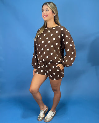 A woman modeling a brown and white polka dot lounge set consisting of a long-sleeve sweatshirt and matching shorts, standing against a solid blue studio backdrop.