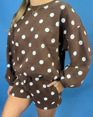 A person wearing a brown long-sleeve sweatshirt and matching shorts set with a white polka dot pattern, posed against a solid blue background.