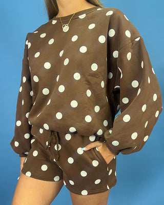 A person wearing a brown long-sleeve sweatshirt and matching shorts set with large white polka dots, photographed against a solid blue background.