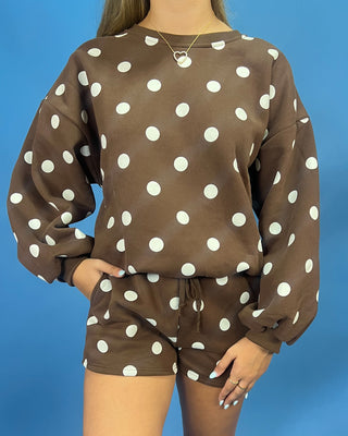 A woman wearing a brown and white polka dot lounge set, featuring a long-sleeve sweatshirt and matching shorts, set against a solid blue background.