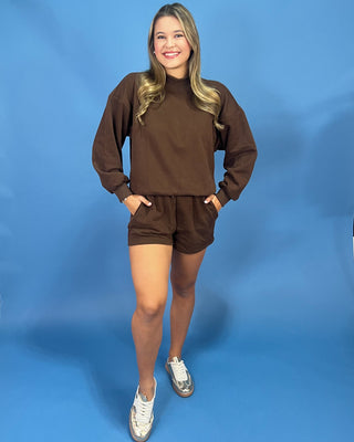 A woman modeling a matching brown lounge set consisting of a long-sleeve crewneck sweatshirt and shorts, paired with white sneakers, against a solid blue studio backdrop.