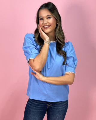 Sky blue short sleeve blouse with puff sleeves and delicate lace trim at the neckline and sleeves, paired with dark wash skinny jeans and metallic thong heels, styled against a pink backdrop.