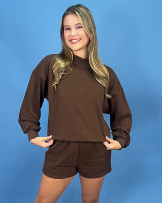 a matching brown long-sleeve mock neck sweatshirt and shorts set, posing against a solid blue studio background.