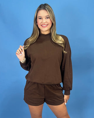 A woman modeling a matching brown long-sleeve mock neck sweatshirt and shorts set against a solid blue studio background.