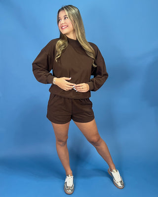 A woman modeling a matching brown lounge set consisting of a long-sleeve sweatshirt and shorts, paired with white sneakers, against a solid blue studio backdrop.