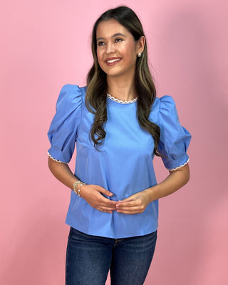 Sky blue short sleeve blouse with puff sleeves and delicate lace trim at the neckline and sleeves, paired with dark wash skinny jeans and metallic thong heels, styled against a pink backdrop.