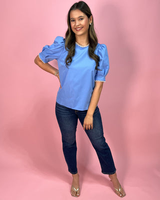 Sky blue short sleeve blouse with puff sleeves and delicate lace trim at the neckline and sleeves, paired with dark wash skinny jeans and metallic thong heels, styled against a pink backdrop.