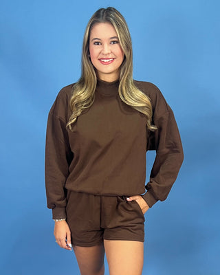 A woman modeling a matching brown long-sleeve mock neck sweatshirt and shorts set against a solid blue studio background.