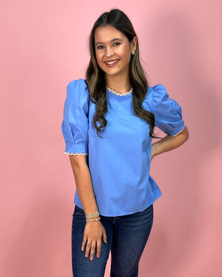 Sky blue short sleeve blouse with puff sleeves and delicate lace trim at the neckline and sleeves, paired with dark wash skinny jeans and metallic thong heels, styled against a pink backdrop.