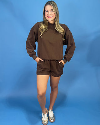 A woman modeling a matching brown sweatshirt and shorts set against a solid blue studio backdrop.