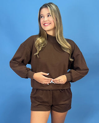 A woman modeling a matching brown sweatshirt and shorts set against a solid blue studio background.
