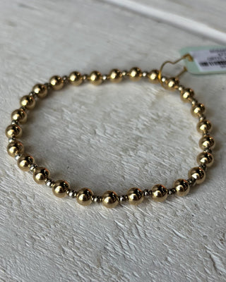 Gold beaded bracelet on a light gray surface with a tag in the background.