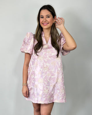 Smiling woman standing against a neutral background wearing a light pink floral mini dress with puff sleeves, styled with nude heeled sandals.