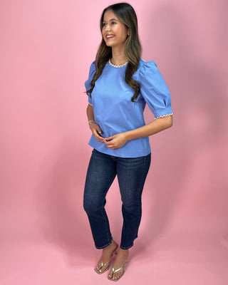 Sky blue short sleeve blouse with puff sleeves and delicate lace trim at the neckline and sleeves, paired with dark wash skinny jeans and metallic thong heels, styled against a pink backdrop.