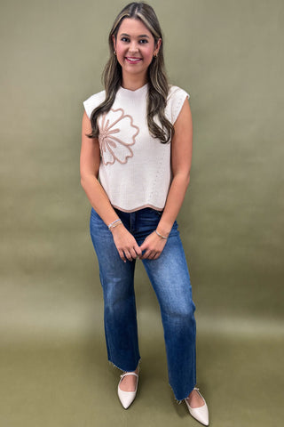 Woman wearing a white sleeveless top with a floral design and blue jeans against a green background