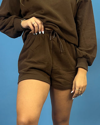 A person wearing a matching brown lounge set consisting of a long-sleeve sweatshirt and drawstring shorts, shown from the chest down against a solid blue background.