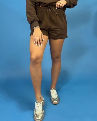A person wearing brown shorts and a matching brown sweatshirt, paired with metallic silver high-top sneakers, standing against a solid blue background.