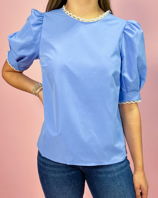 Sky blue short sleeve blouse with puff sleeves and delicate lace trim at the neckline and sleeves, paired with dark wash skinny jeans and metallic thong heels, styled against a pink backdrop.