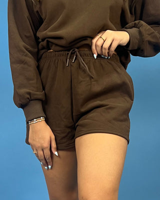 A person wearing a matching brown sweatshirt and shorts set, shown from the chest down against a solid blue background.