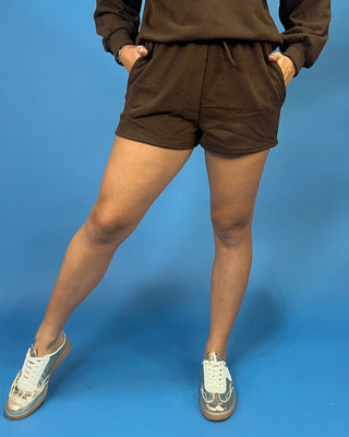 A person wearing brown drawstring shorts and a matching brown sweatshirt, standing against a solid blue studio background with white sneakers.