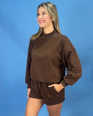A woman modeling a brown long-sleeve mock neck sweatshirt and matching brown shorts against a solid blue studio background.