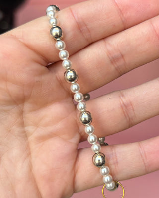 Hand holding a bracelet with alternating pearls and metallic beads against a pink background