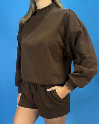 A person wearing a dark brown long-sleeve mock neck sweatshirt and matching dark brown shorts, standing against a solid blue background.