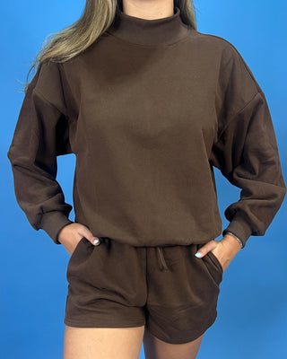 A person wearing a matching brown long-sleeve mock-neck sweatshirt and shorts set, standing against a solid blue background with hands in the shorts pockets.