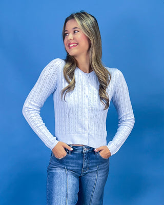 A woman with blonde-highlighted hair wearing a light blue ribbed knit cardigan and blue jeans, posing against a solid blue studio background.
