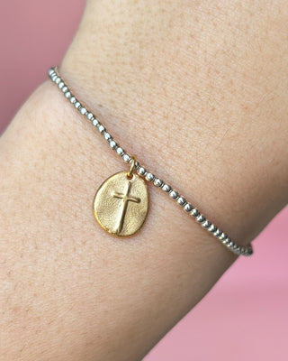 Gold bracelet with a cross charm on a wrist against a pink background