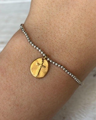 Gold bracelet with a cross charm on a wrist against a neutral background