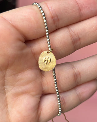 Gold charm on a beaded bracelet held in a hand with a pink background