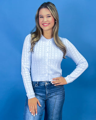 A woman modeling a light blue ribbed knit cardigan with button-front closure, paired with blue jeans, against a solid blue studio background.