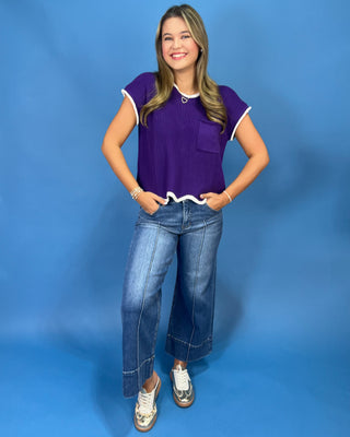 A woman modeling a purple short-sleeve ribbed knit top with white trim, paired with blue jeans and white sneakers, standing against a solid blue studio backdrop.